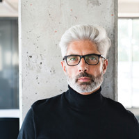 Portrait of a man with silver hair and glasses in a black turtleneck.