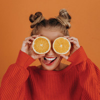Woman holding orange slices over her eyes against an orange background.